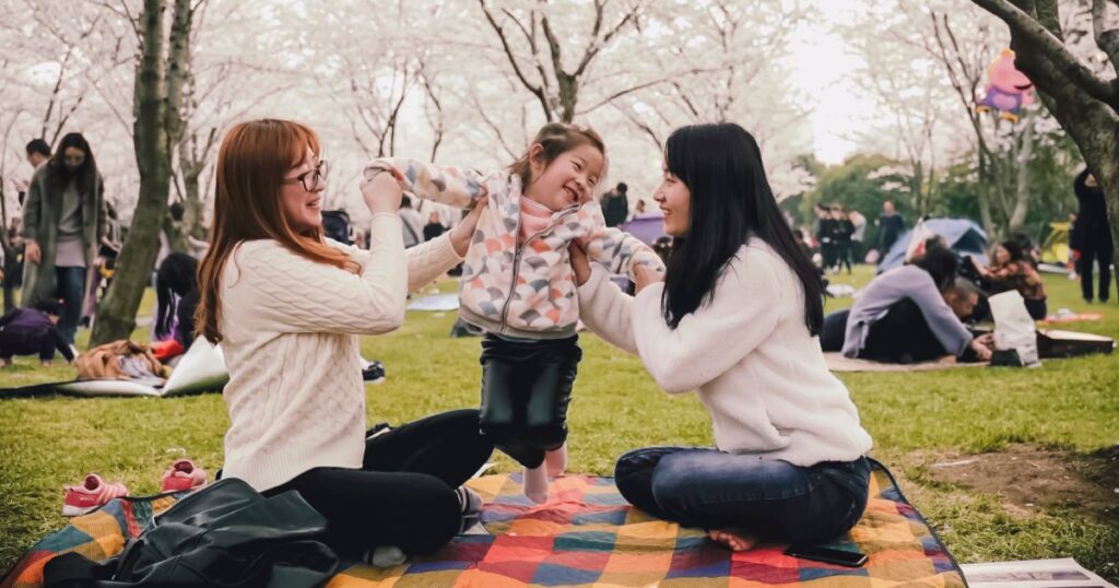 two moms holding laughing child on picnic blanket in park